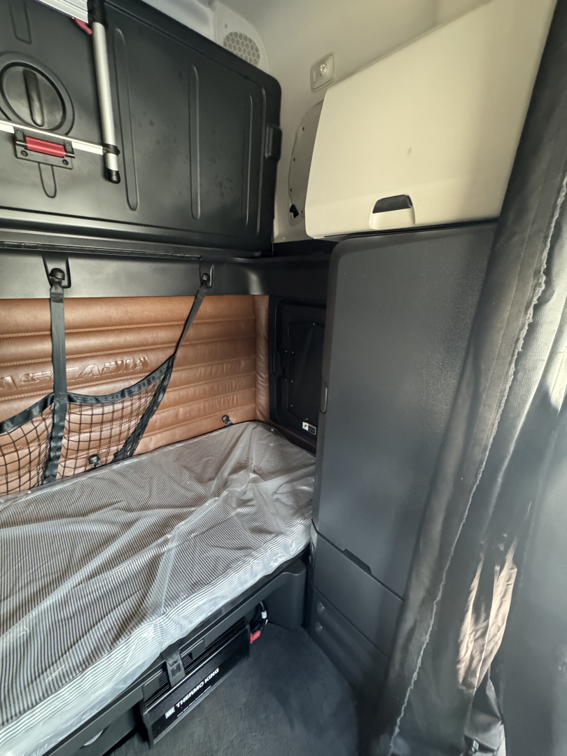 Cab interior of a Freightliner Cascadia 126 sleeper showing bunk bed with gray sheet, brown padded wall, overhead storage, and side cabinet.