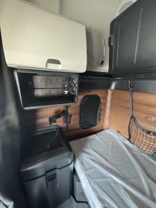 Cab interior of a Freightliner Cascadia 126 sleeper showing side storage, a small shelf, brown leatherette paneling, and a mattress with a light cover.
