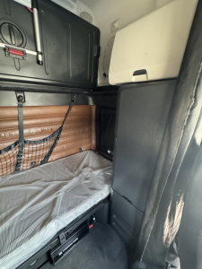 Cab interior of a Freightliner Cascadia 126 sleeper showing bunk bed with gray sheet, brown padded wall, overhead storage, and side cabinet.