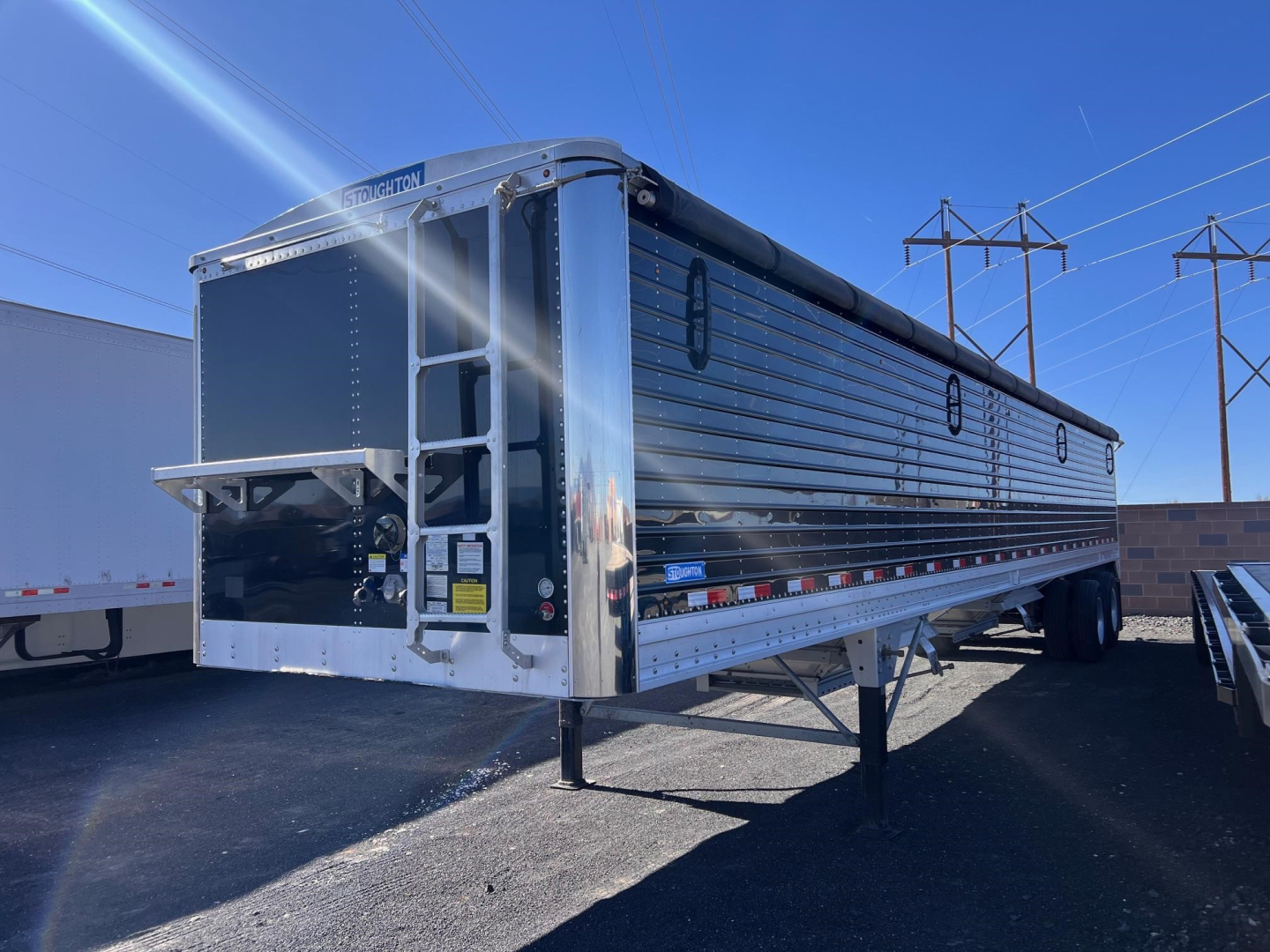 Stoughton grain trailer hopper trailer side and rear with chrome panels and ladders, elevated hitch, in a lot. Visible text includes Stoughton logo and reflective tape along side.