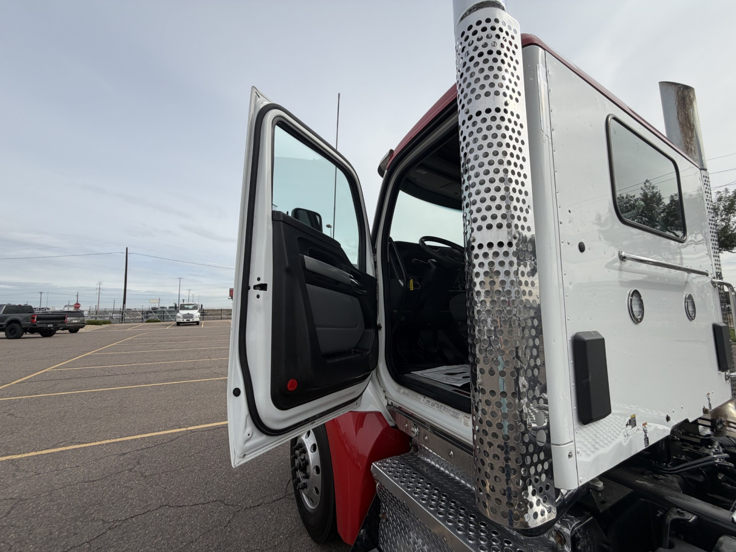 Kenworth T880 cab with open door, side step, and perforated metal exhaust stack panel visible. Vehicle appears white with red accents and is in a parking lot.