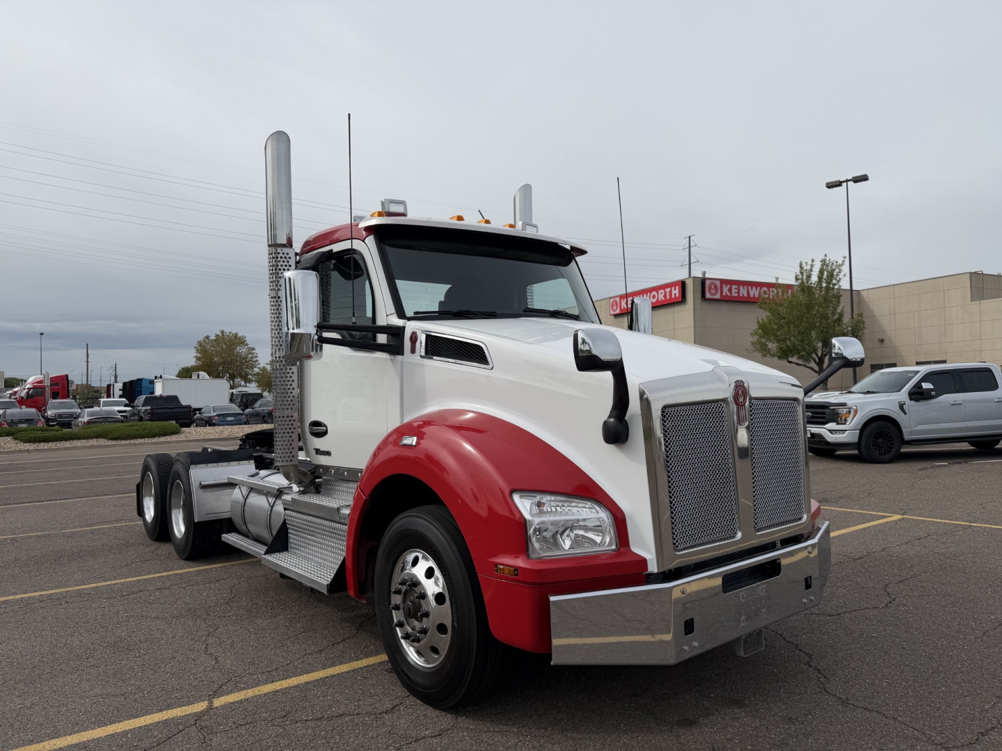 Kenworth T880 conventional daycab truck in white and red, front grille, dual vertical exhaust stacks, chrome bumper, daytime running lights, parked in lot.