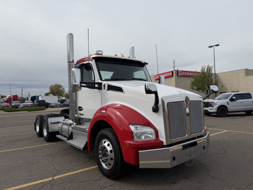 Kenworth T880 conventional daycab truck in white and red, front grille, dual vertical exhaust stacks, chrome bumper, daytime running lights, parked in lot.