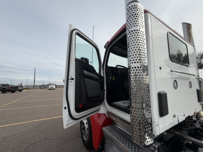 Kenworth T880 cab with open door, side step, and perforated metal exhaust stack panel visible. Vehicle appears white with red accents and is in a parking lot.