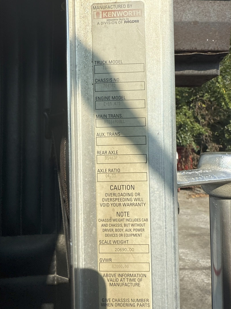 Kenworth truck chassis information label on a vertical surface showing manufactured by Kenworth, truck model, chassis number, engine model, rear axle, axle ratio, gross vehicle weight rating (GVWR) and cautions.