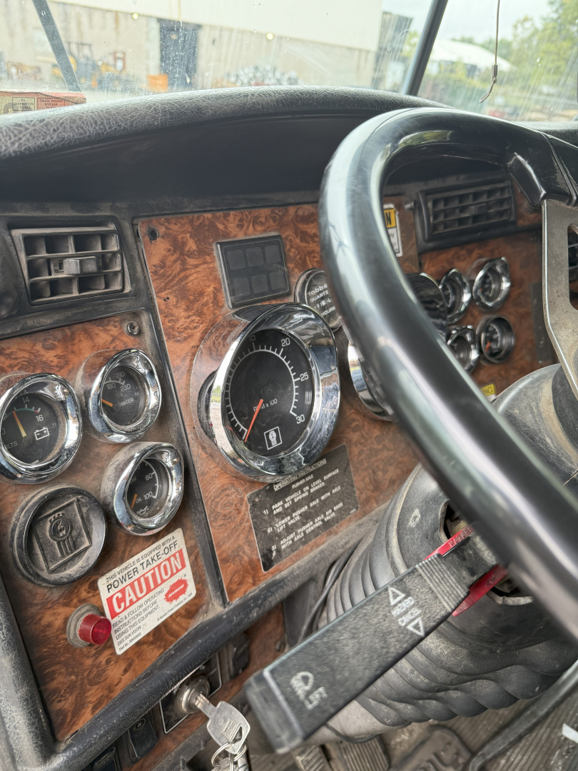 Truck dashboard showing woodgrain panel, analog gauges, steering wheel, and control stalks in a Kenworth T800 cab interior.