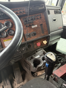 Truck interior dash of Kenworth T800 showing wood-grain panel, steering wheel, gauge cluster, floor pedals, shifter, and gear lever.