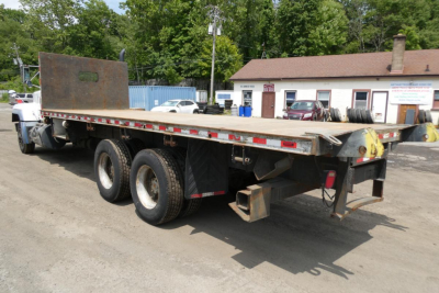 Flatbed truck trailer mounted on a Mack RD690S chassis showing dual rear axles and a rusted storage plate at the front, side rails with reflective tape.