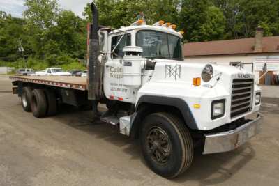 Flatbed tow truck showing Mack RD690S front view with white cab and long flatbed, text on door, and orange cab lights.