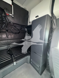 Cab interior of Freightliner Cascadia showing storage cabinet and sleeper area with seat and panels.