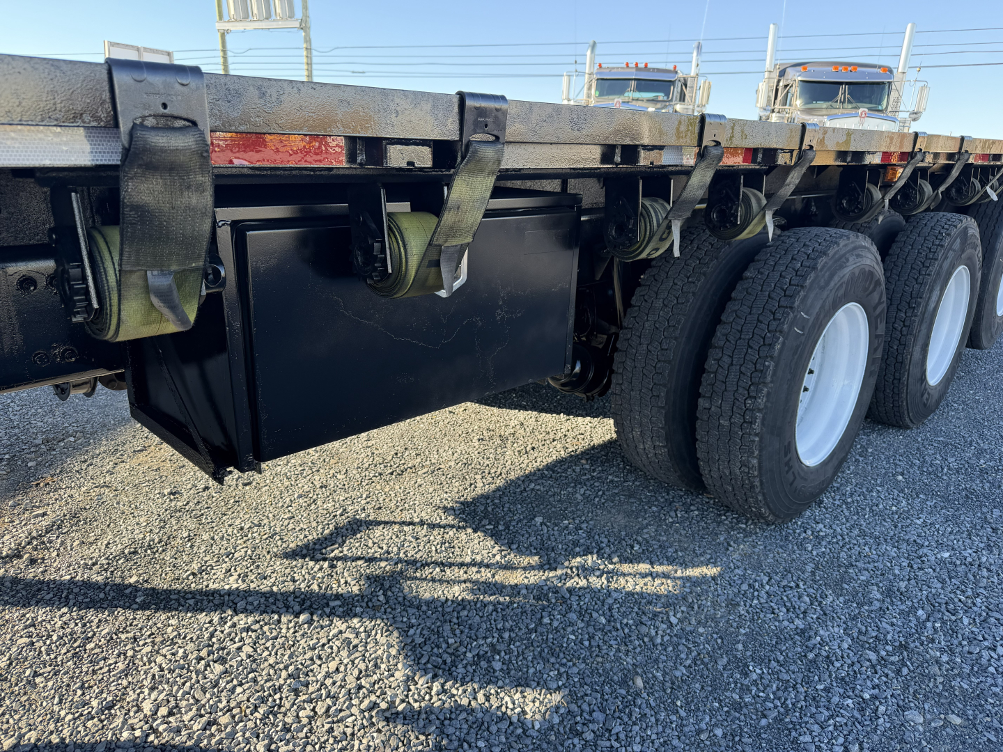 Flatbed trailer undercarriage showing black tool/haul storage box and dual-axle wheels with white rims, near-ground gravel surface.