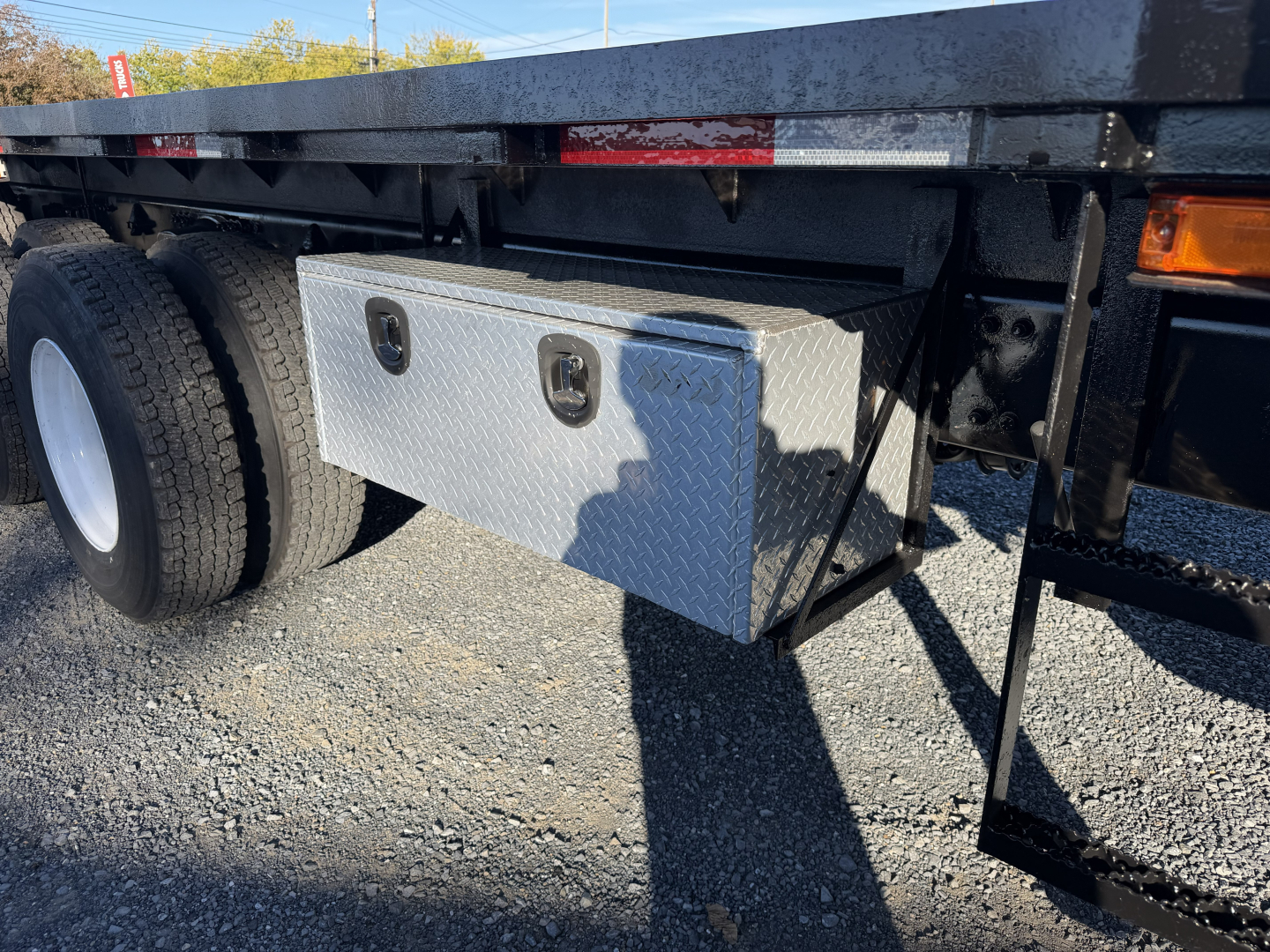 Flatbed truck undercarriage showing aluminum tool/storage toolbox mounted to frame with dual louvered access eyes, large rear wheels, and metallic steps.