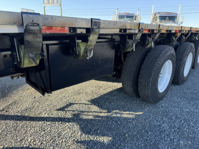Flatbed trailer undercarriage showing black tool/haul storage box and dual-axle wheels with white rims, near-ground gravel surface.