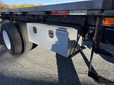 Flatbed truck undercarriage showing aluminum tool/storage toolbox mounted to frame with dual louvered access eyes, large rear wheels, and metallic steps.