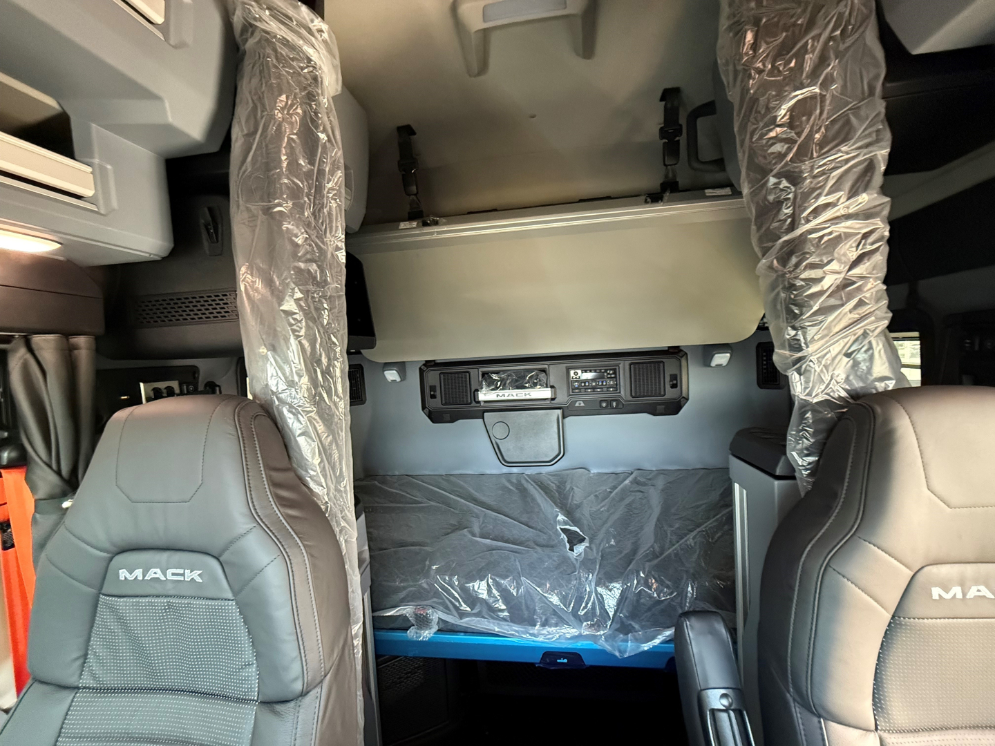 Cab interior of a 2027 Mack AN64T64 truck showing front seats with protective plastic, overhead console, and bunk area in the rear, dash controls visible.