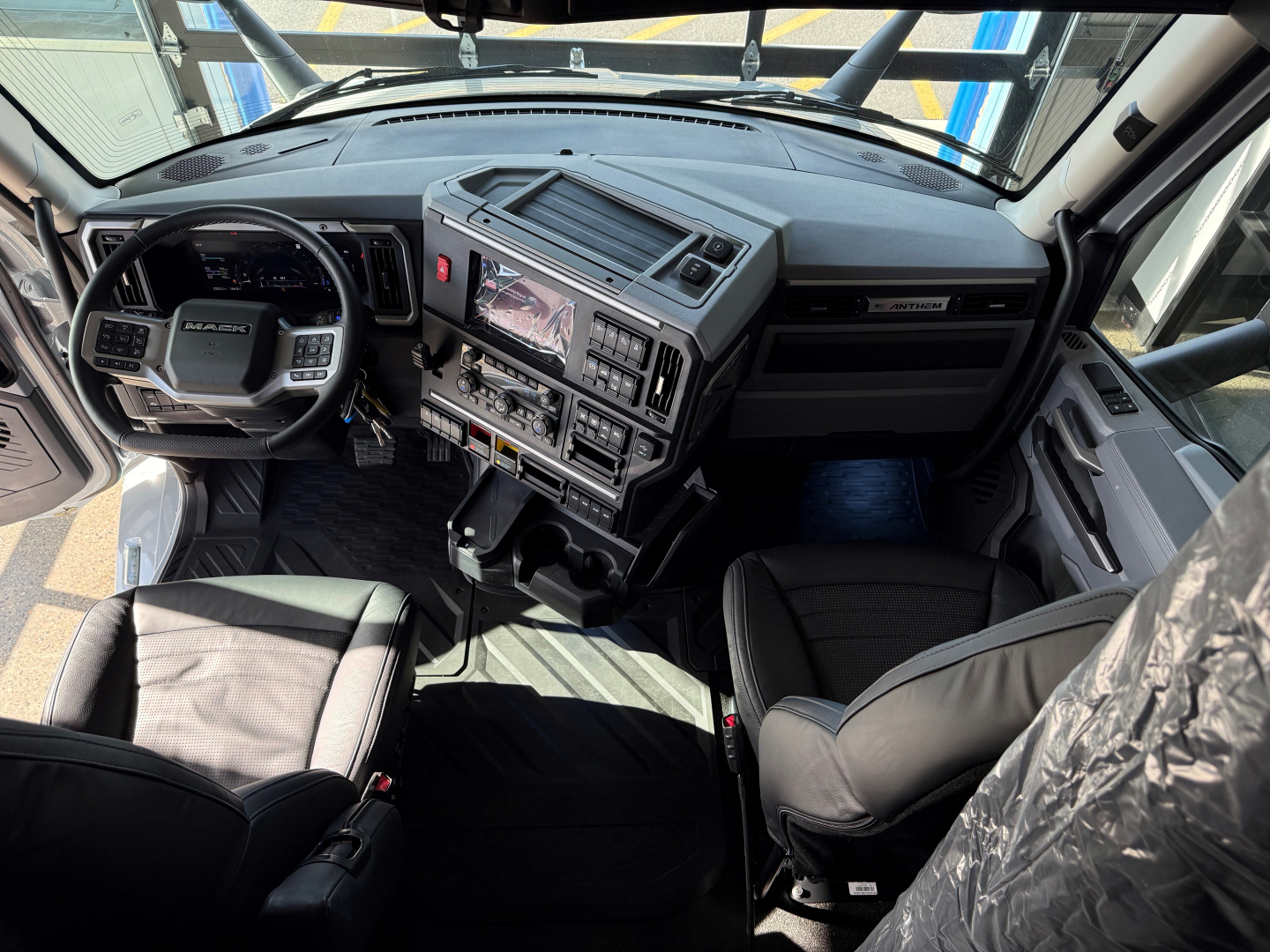 Cockpit view of a Mack truck interior showing dashboard with touchscreen display, steering wheel, and driver and passenger seats.