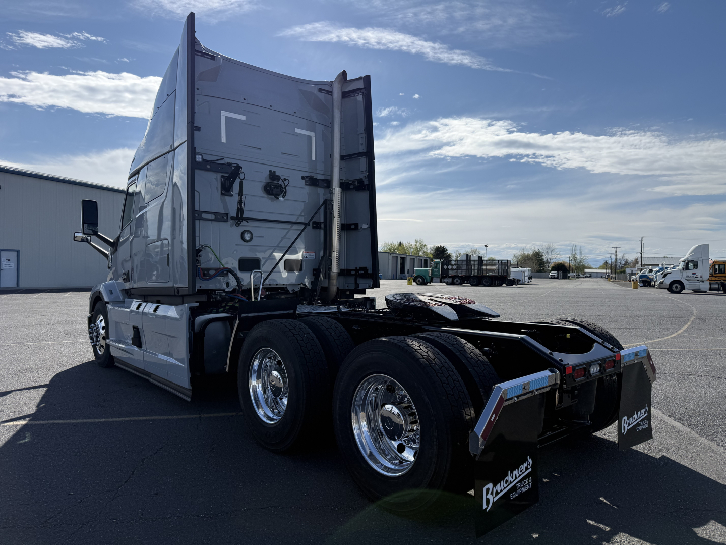 Truck chassis with rear view showing tandem axles and fifth wheel mount. Visible text includes Mack AN64T64 branding on mudflaps and Brucker? dealership sticker on rear. Clear blue sky.