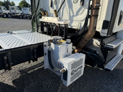 Truck engine bay area of a Freightliner Cascadia 125DC showing auxiliary fuel or air system with metal braided hoses and mounting plate.