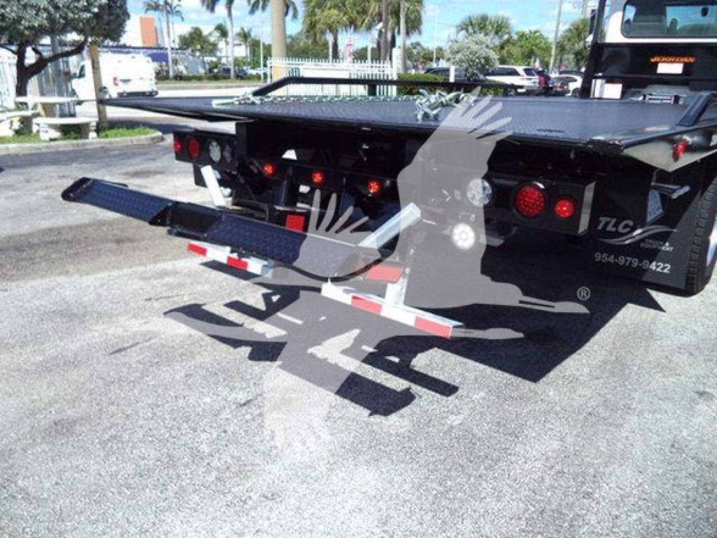 Rear view of a rollback tow truck with a mounted flatbed and hydraulic lift, showing taillights, rear bumper, and TLC logo on the side.