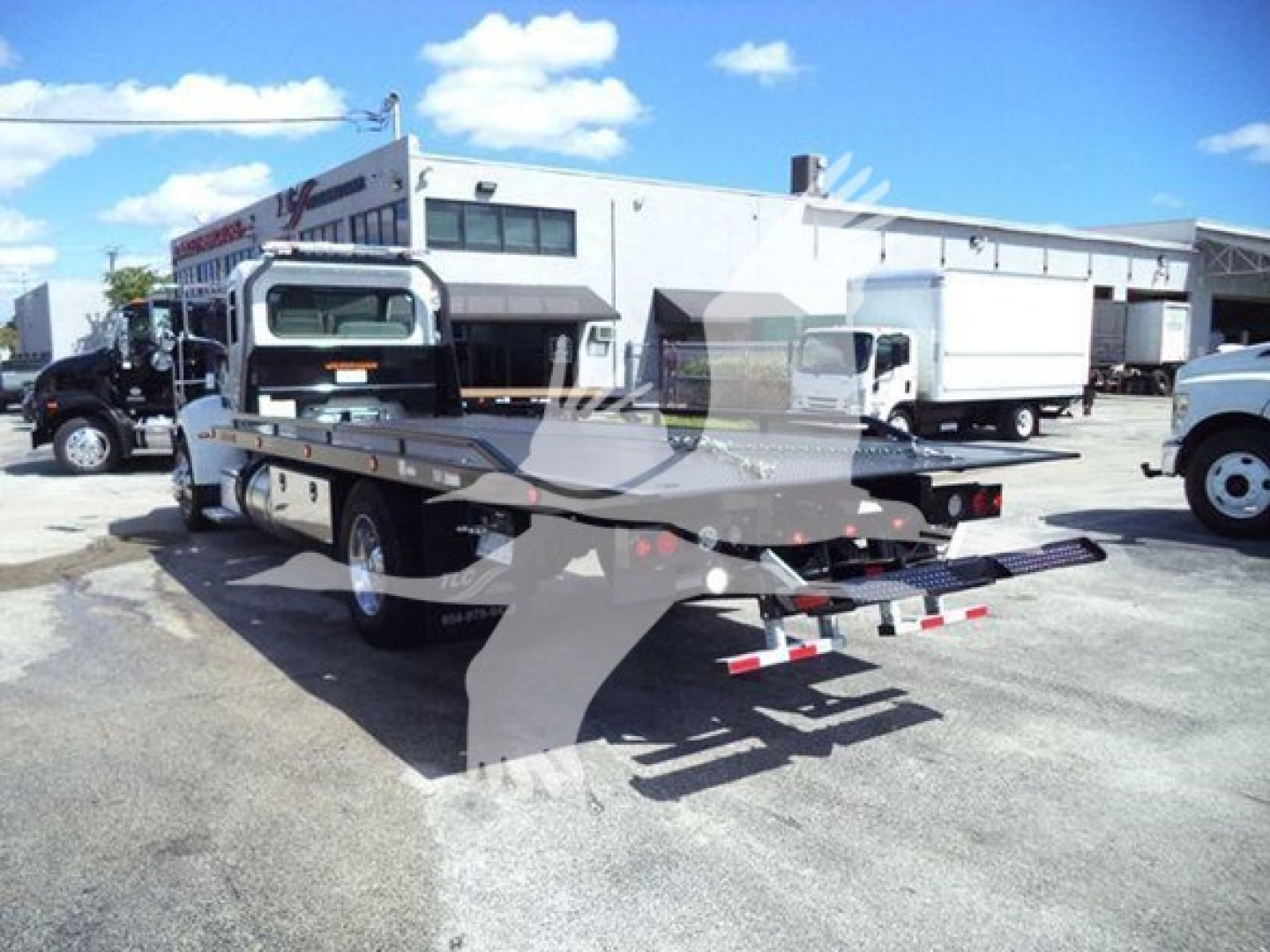 Peterbilt 337 rollback tow truck with flatbed, side view, rear apparatus visible; yard with buildings and other trucks in background.