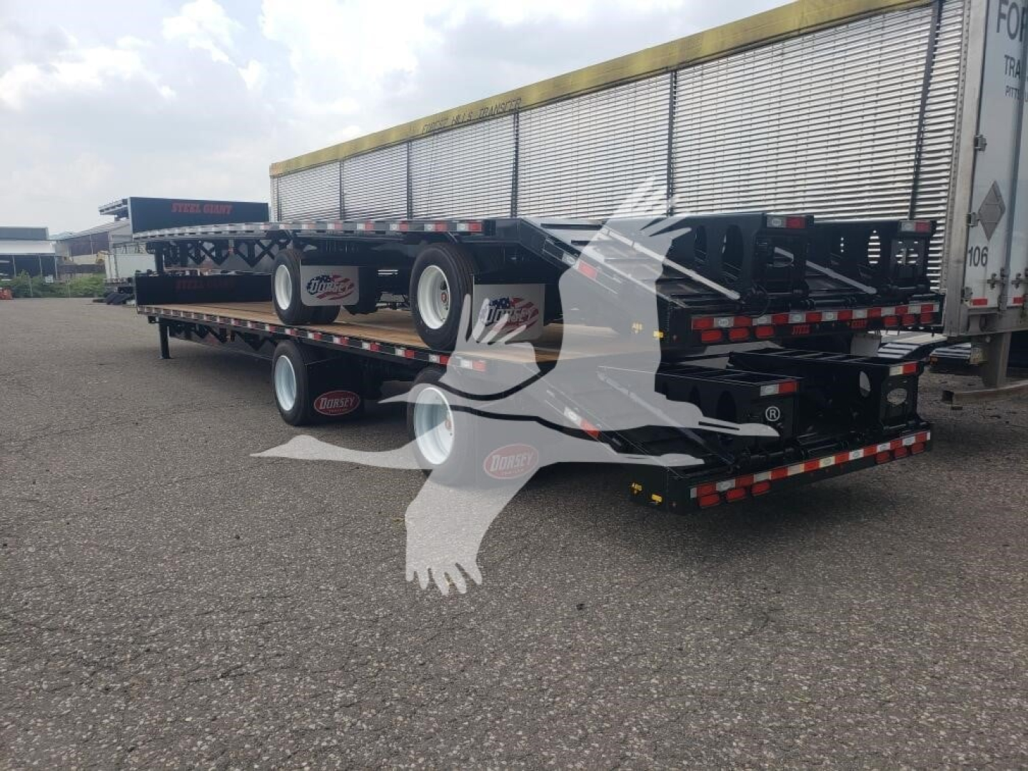 Trailer deck with beavertail trailer in a parking lot, rear view showing multiple axles and flatbed sections.