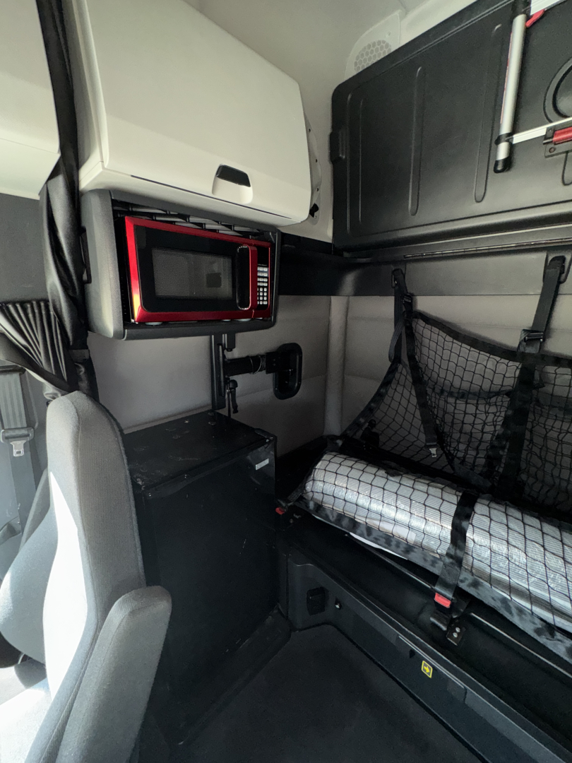 Cab interior of a 2021 Freightliner Cascadia 126 sleeper showing a mounted microwave, storage cabinet, and seating area with a protective net.
