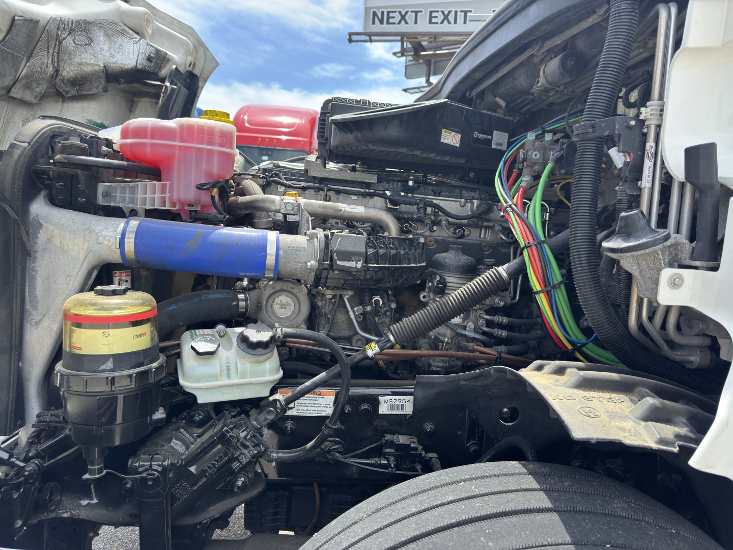 Freightliner Cascadia engine bay showing coolant reservoir, pink coolant overflow, blue coolant hose, multiple colored electrical harnesses, and various fluids/filters.