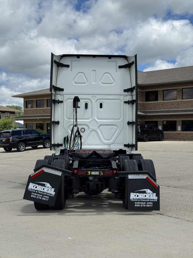 Rear view of a Freightliner truck tractor chassis with fifth wheel removed, showing back of cab and truck mud flaps with dealership branding.