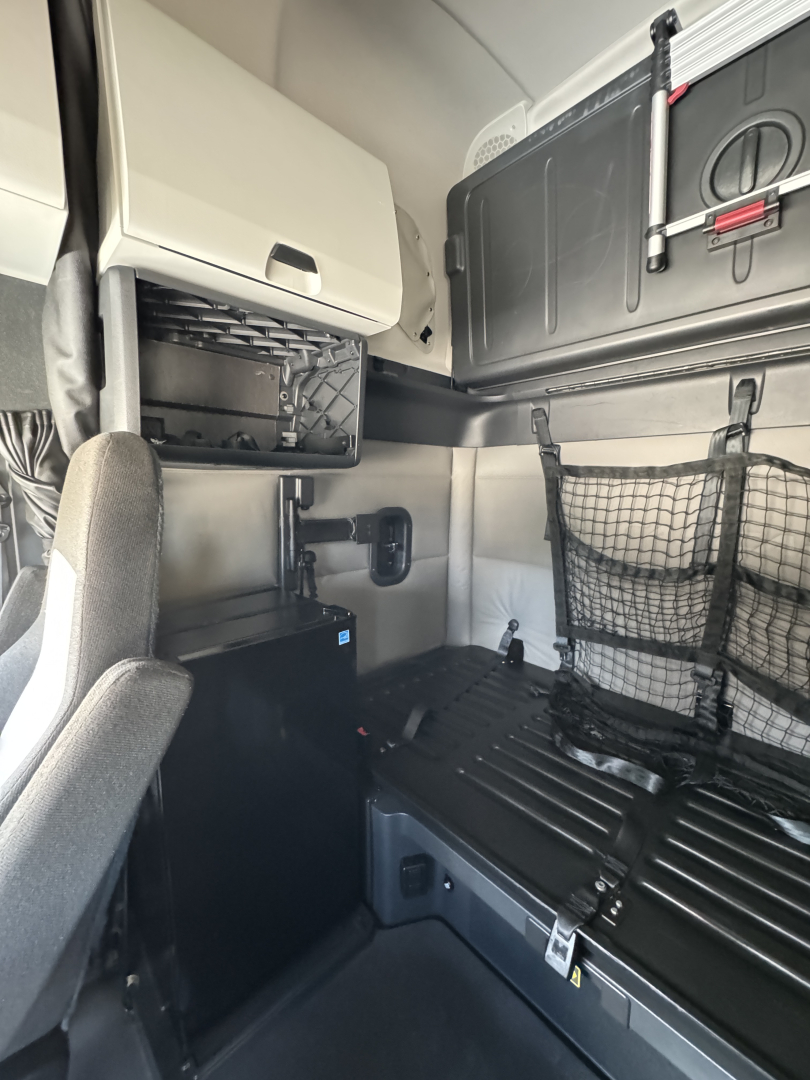 Front cab interior of a Freightliner Cascadia sleeper showing storage shelf, overhead cabinet, nets, and seating area.
