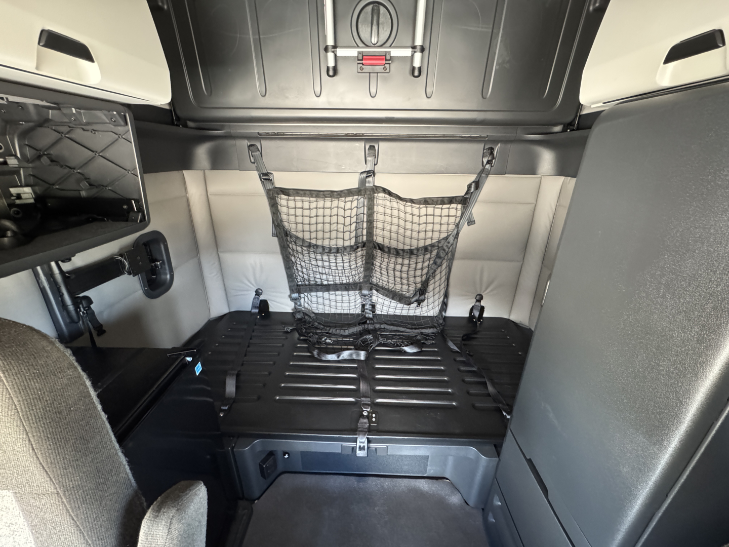 Truck cab interior showing storage net on back wall and black plastic cargo area in Freightliner Cascadia sleeper, with metallic walls and overhead panels.