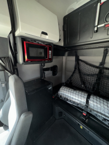 Cab interior of a 2021 Freightliner Cascadia 126 sleeper showing a mounted microwave, storage cabinet, and seating area with a protective net.
