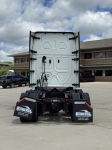 Rear view of a Freightliner truck tractor chassis with fifth wheel removed, showing back of cab and truck mud flaps with dealership branding.
