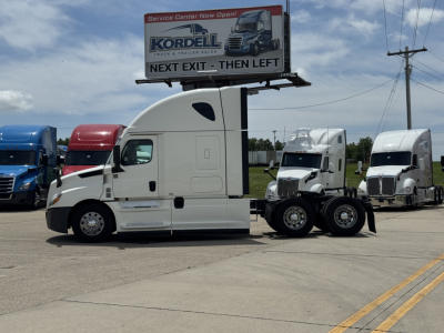 Freightliner Cascadia 126 semi-truck tractor in white, side view, with two matching trucks in background and a billboard.