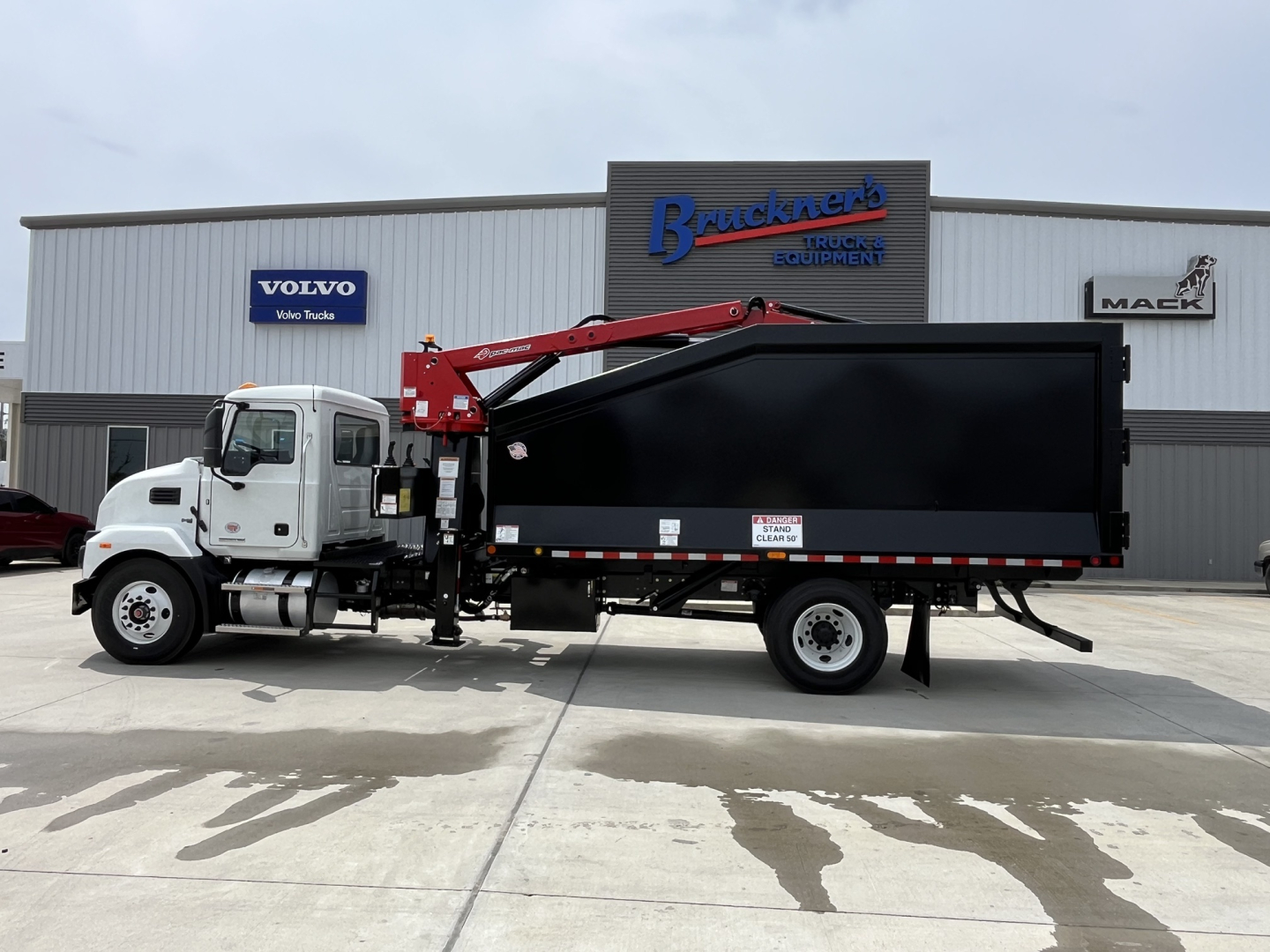 New 2025 Mack MD7 grapple truck with a black dump body and red hydraulic grapple arm, side view in front of dealership building signage.