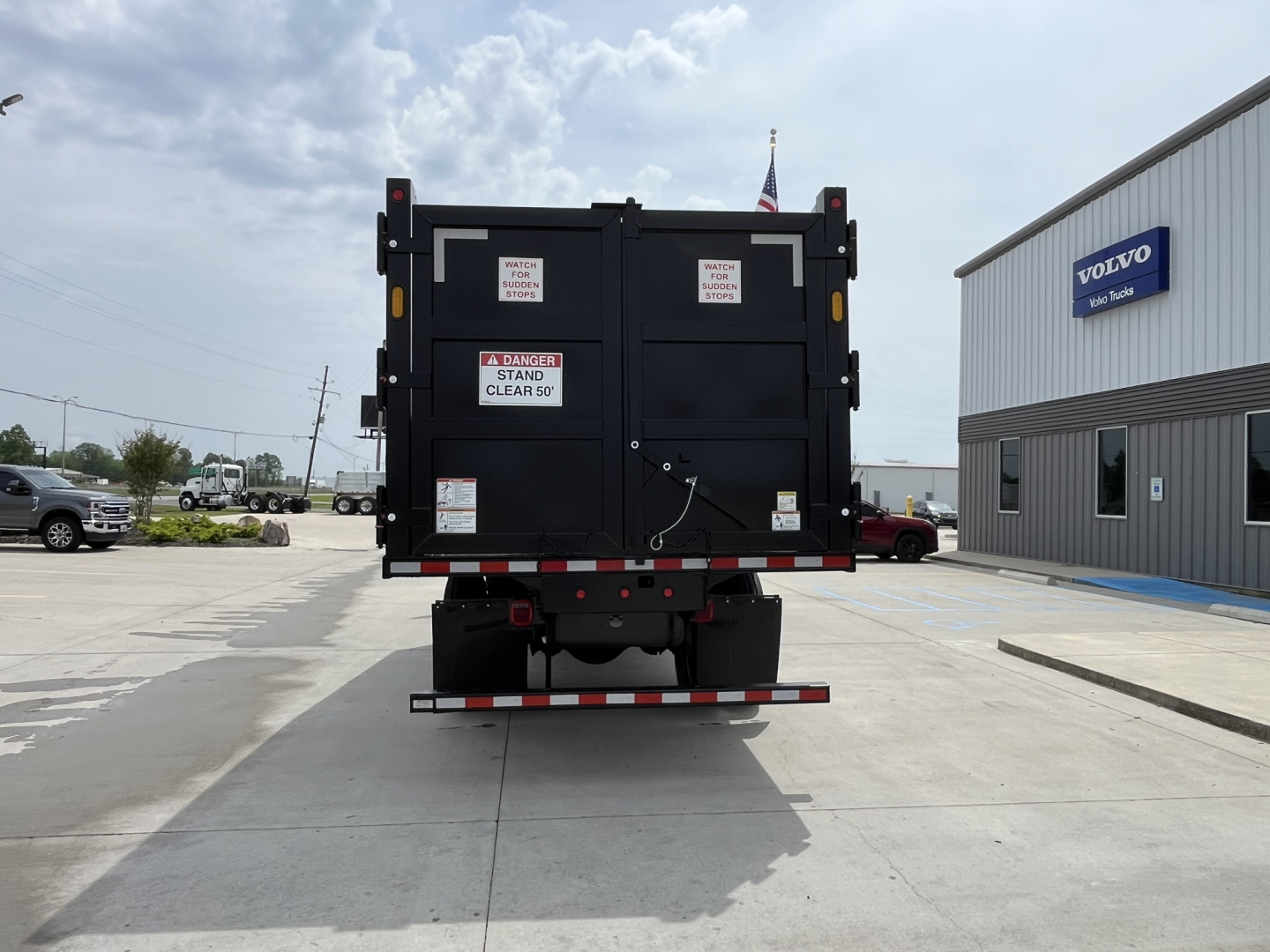 Back view of new 2025 Mack MD7 grapple truck with rear doors, safety stickers, and high-visibility markings in a dealership lot.