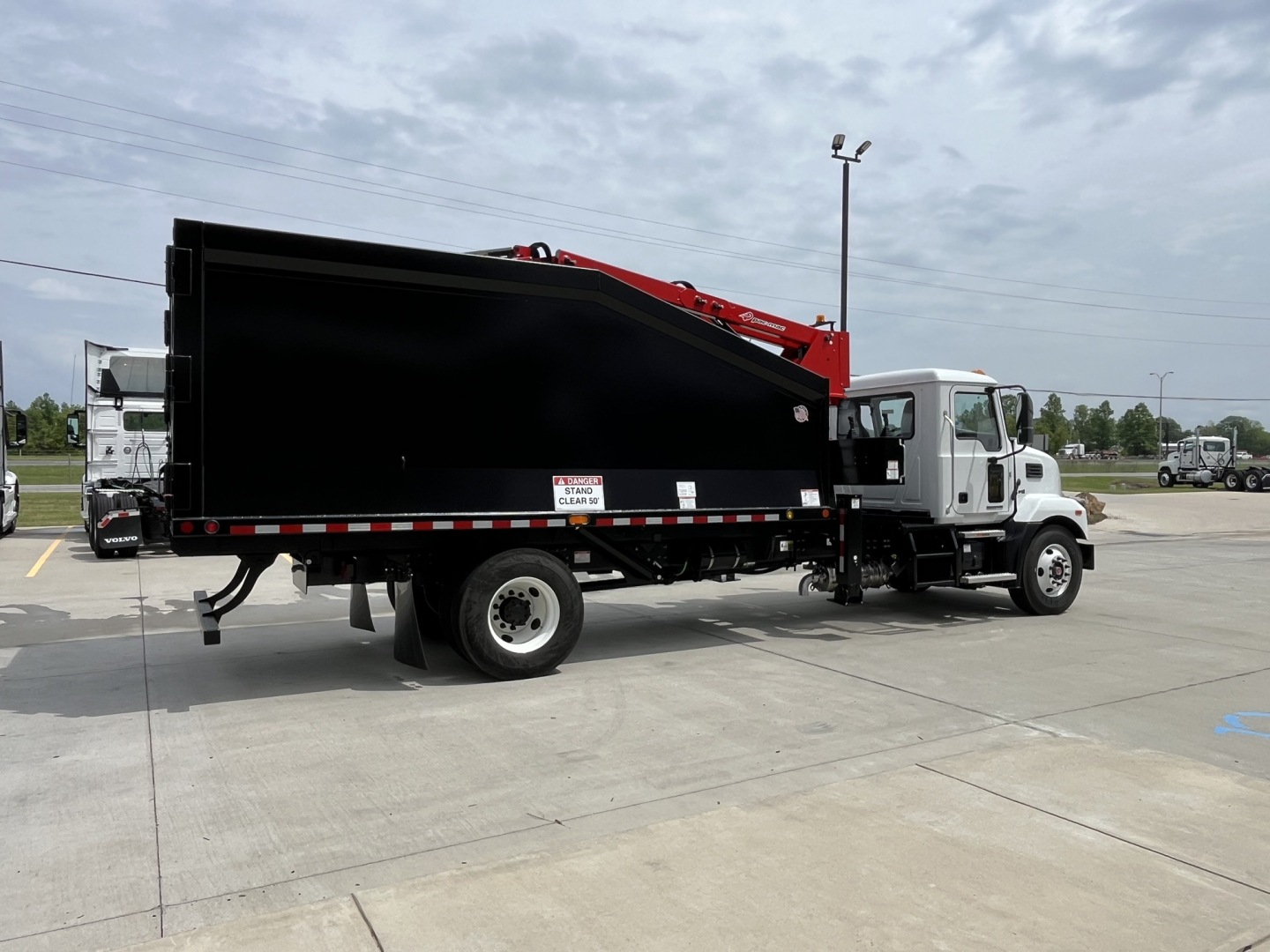 Truck chassis with a large black dump bed and a red crane attached, Mack MD7 in background, side view showing cab and dump body.