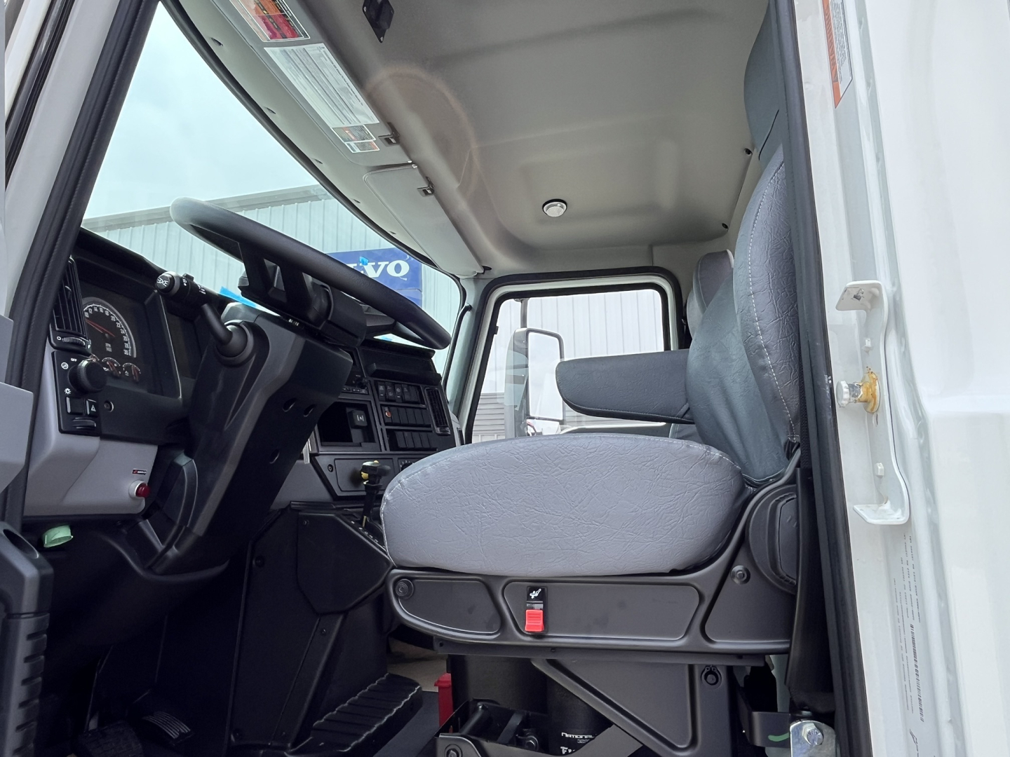 Cab interior of a 2025 Mack MD7 truck showing driver seat and dashboard from open door. Visible steering wheel, instrument cluster, pedals, and passenger seat area.