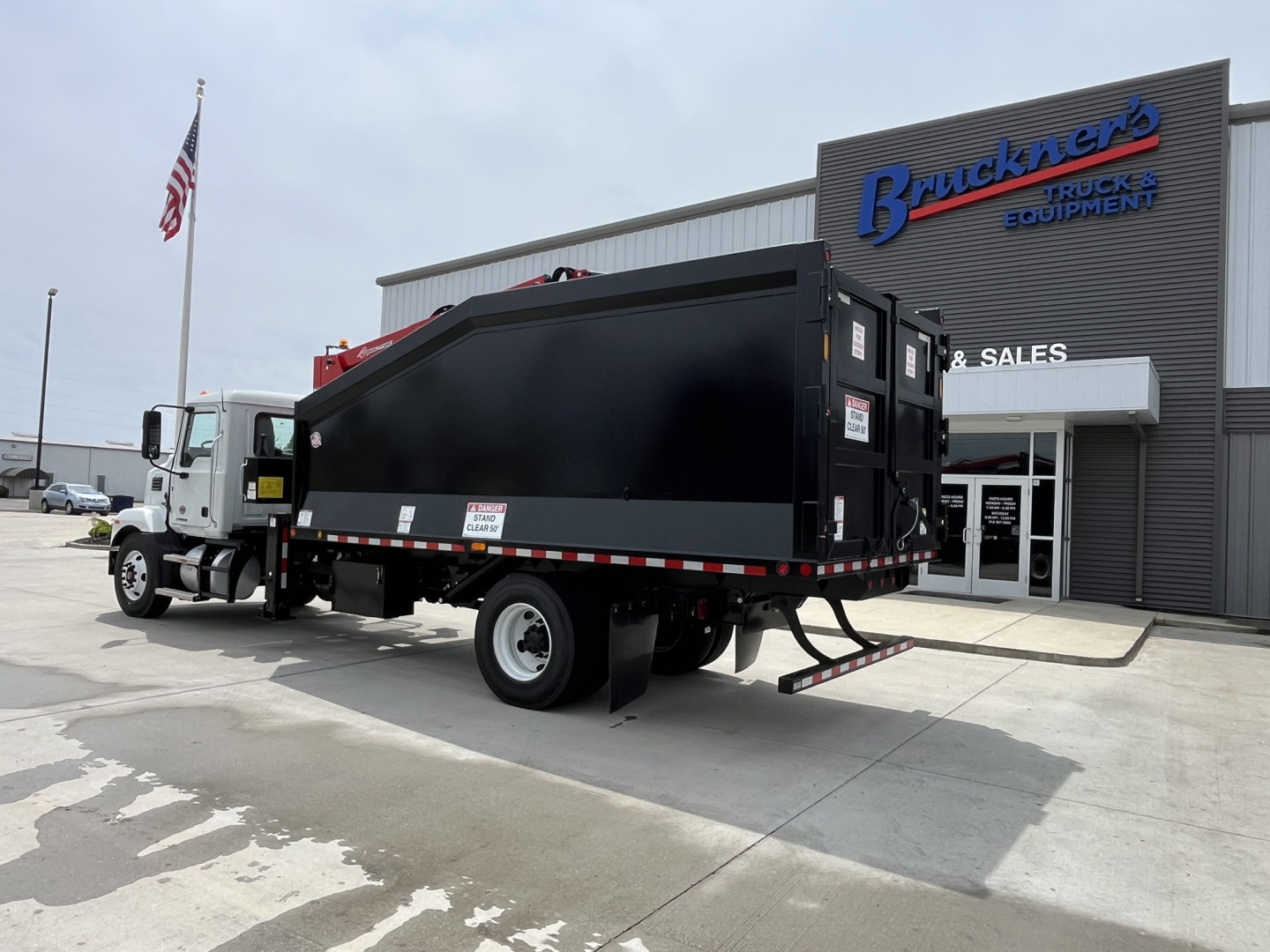 Truck with a black dump body in front of a dealership, showing a white cab and a large black grapple/dump bed mounted on a Mack MD7 chassis.
