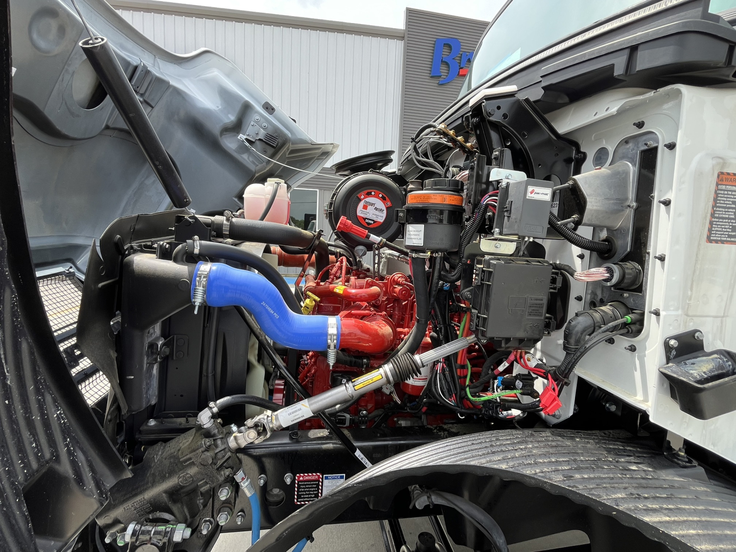 Mack MD7 engine bay showing red and blue hoses, belts, and electrical components under open hood; visible gauges and wiring harnesses.