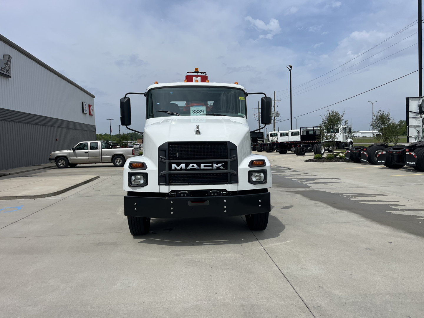 Front view of a white Mack MD7 truck with black grille and bumper, showing the Mack logo; side mirrors; red beacon on roof; other trucks in background.
