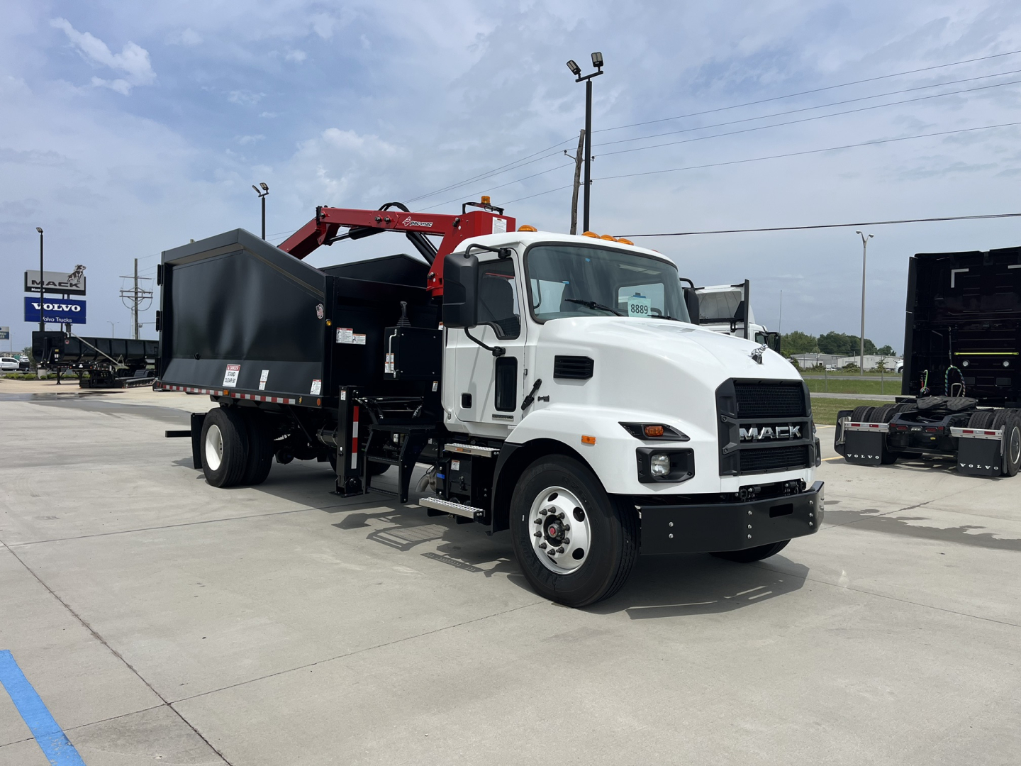 Mack MD7 truck with grapple body in a dealership lot showing white cab and red grapple arm attached to black dump body.
