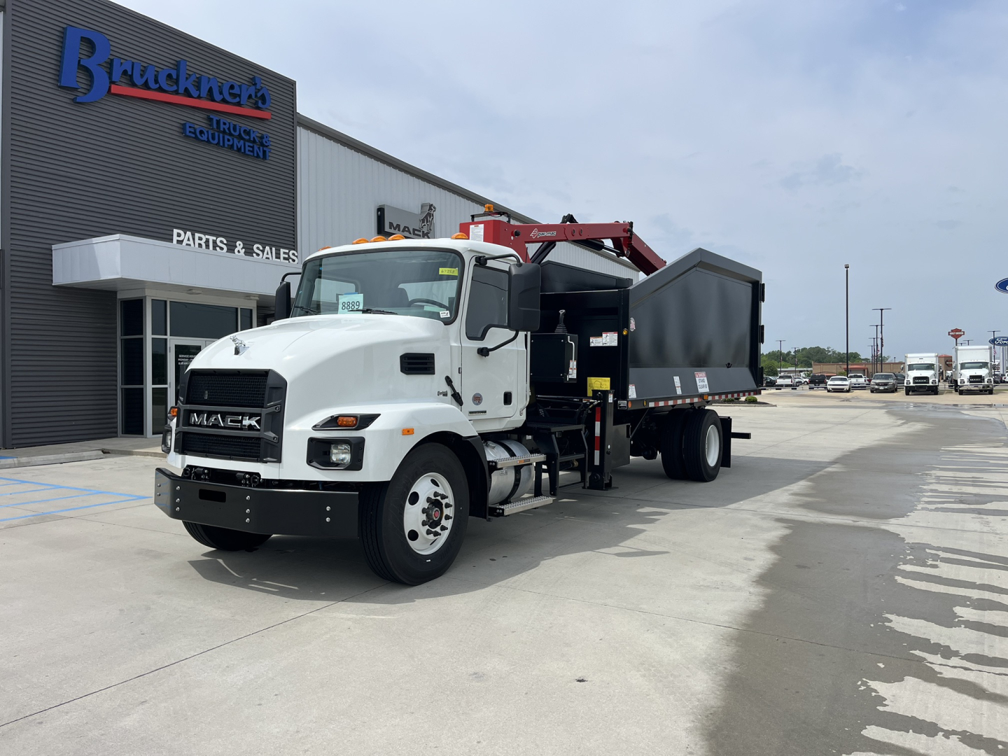 Mack MD7 truck with a grapple/dump body in a dealership lot showing white cab and black bed.