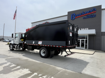 Truck with a black dump body in front of a dealership, showing a white cab and a large black grapple/dump bed mounted on a Mack MD7 chassis.