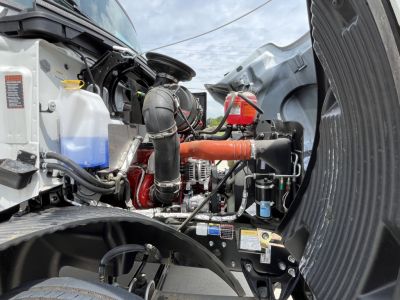 Mack MD7 engine bay with label-covered components, hoses, and orange pipe; visible coolant reservoir, air intake, and electrical wiring in a truck chassis.