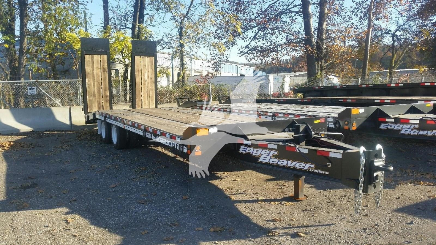 Trailer deck with dual ramps and black and white Eager Beaver branding; visible chains and hitch in foreground; trees and other trailers in background.