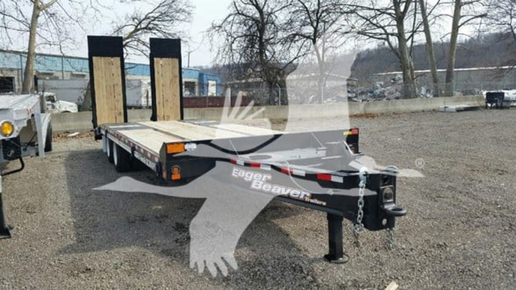 Flatbed trailer showing Eager Beaver logo, dual ramps, black frame, front hitch with safety chains, orange reflectors, gravel lot.