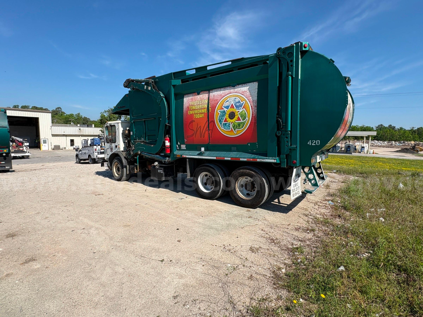 Garbage truck in teal with circular rear tank, side decals, and numbers on rear; Mack MRU613 in yard.