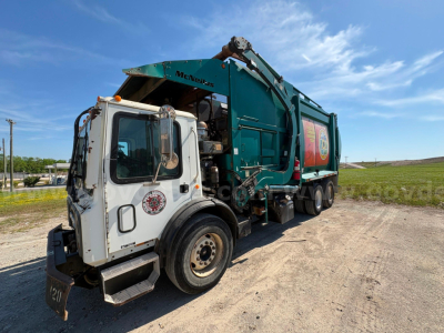 2012 Mack MRU613 garbage truck in a yard, side view, teal refuse body, white cab with logo, front-left wheel visible, hydraulic arm over rear body.