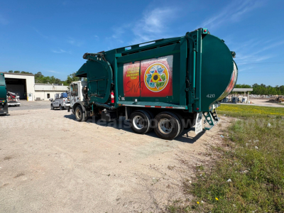 Garbage truck in teal with circular rear tank, side decals, and numbers on rear; Mack MRU613 in yard.