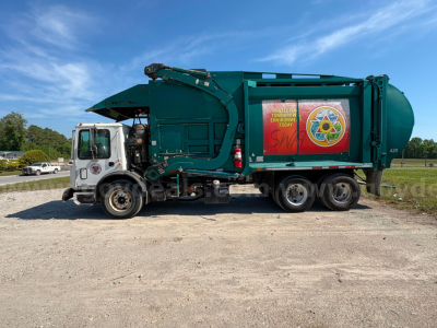 2012 Mack MRU613 garbage truck side view showing waste compactor body and rear wheels; side panel displays graphic with logo and warning text.